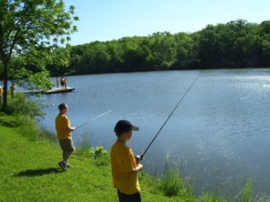 Two young boys fishing on a river, other kids fishing on a dock further in the background